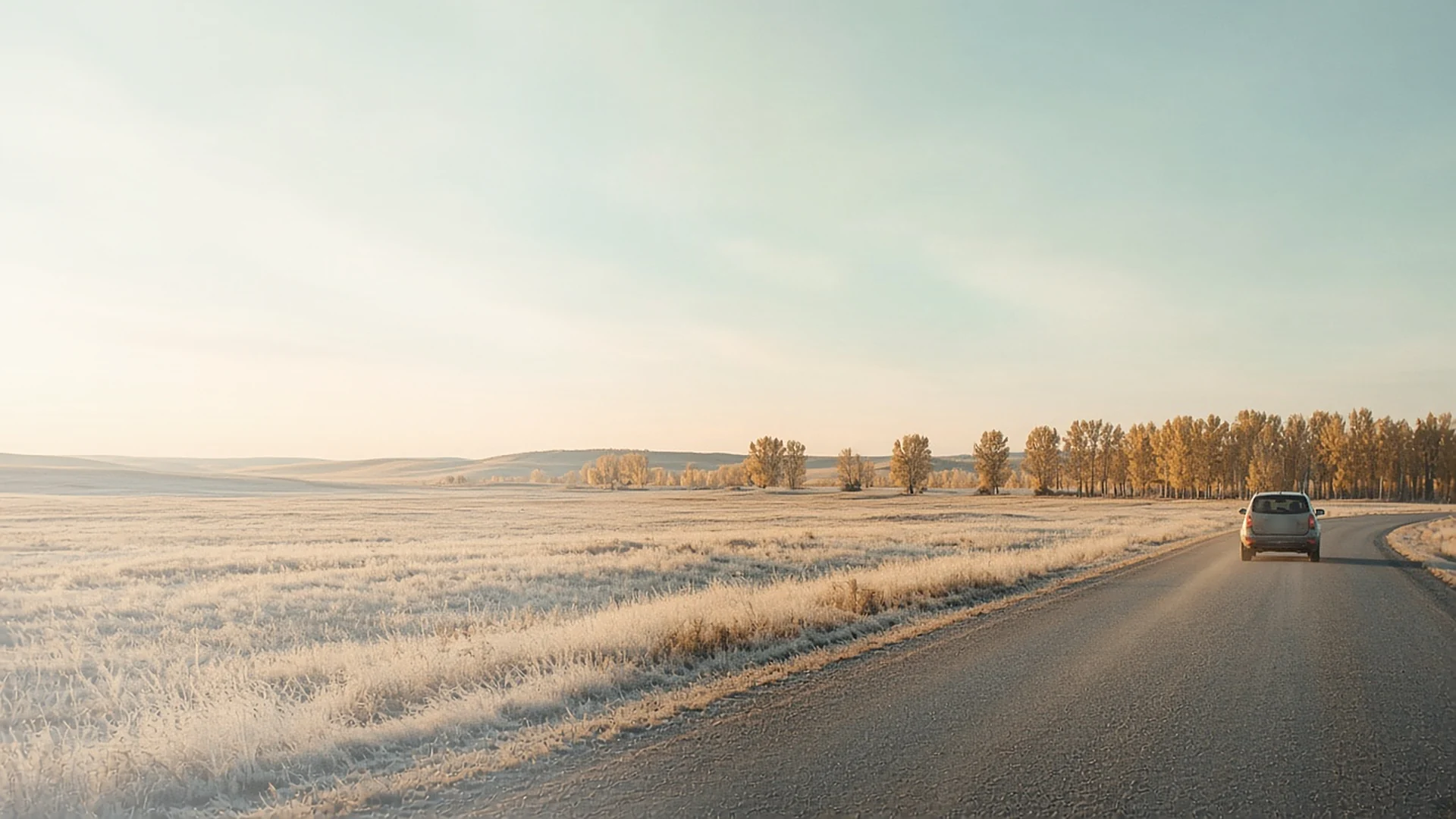 A quiet road through frosted prairie fields at sunrise, with one vehicle in the distance
